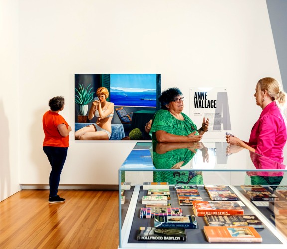 Three people inside a exhibition space. One person is looking at a painting on the wall while the two others are talking. 