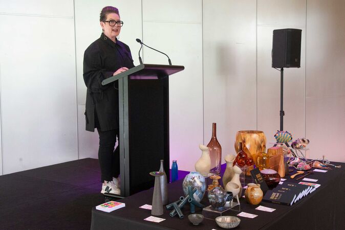 woman in black clothing at speaker podium with display of ceramics and craftwork on table in front of her