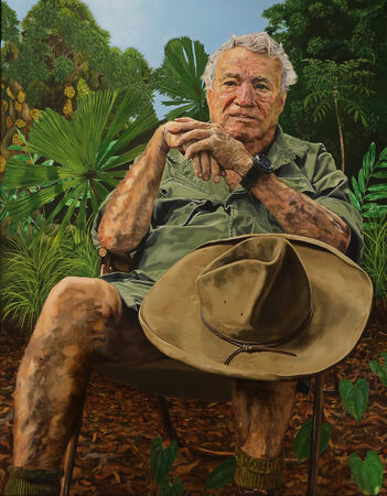 Portrait of an older man in khaki outdoors, seated with his hat on his knee, surrounded by tropical greenery.