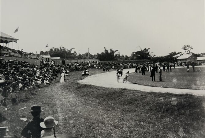 Black-and-white photograph of a cycling race at Mackay Showgrounds, with two riders rounding a curved track. Large crowds sit and stand along grassy embankments and grandstands, many holding umbrellas. Officials and spectators gather near the track, with buildings and trees visible in the background.