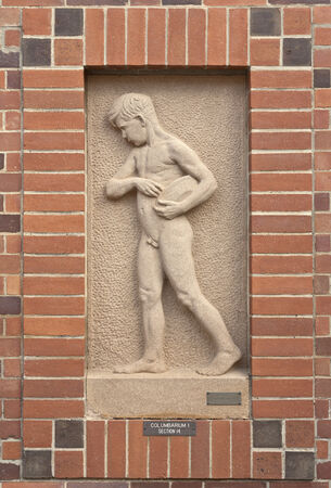 Stone relief sculpture of a nude young male holding an object, set in a textured niche. The frame is made of red and dark brown bricks.