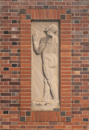A stone relief sculpture of a nude man in profile is set against a textured background, framed by red bricks on a brick wall.