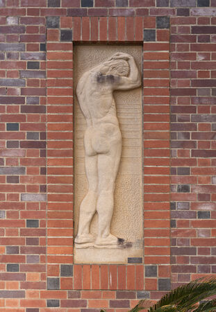 A stone relief of a nude male figure with hands on head is set within a red and dark brick wall.