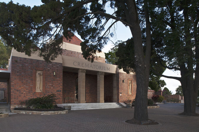 Brick crematorium building with large entrance columns, surrounded by tall trees. The atmosphere is solemn, with an empty paved area in the foreground.