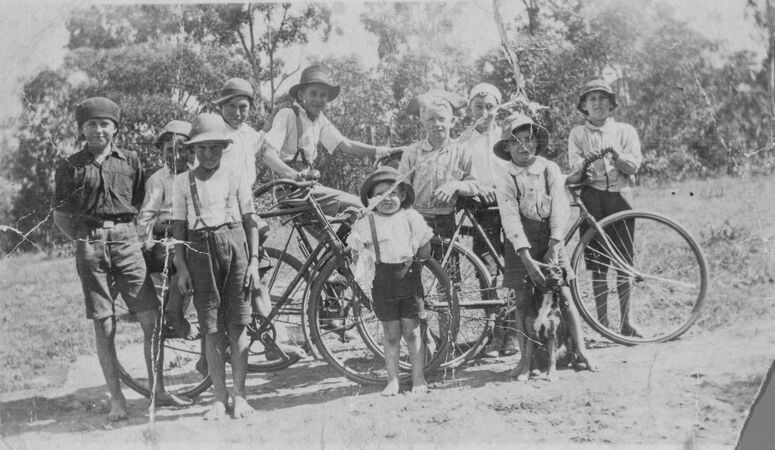 Black-and-white photograph of ten boys outdoors with bicycles, likely from the 1920s. They wear hats, shirts, and suspenders, posing casually on a dirt track with trees behind them. One younger child stands barefoot in front, while others lean on or sit beside their bicycles.