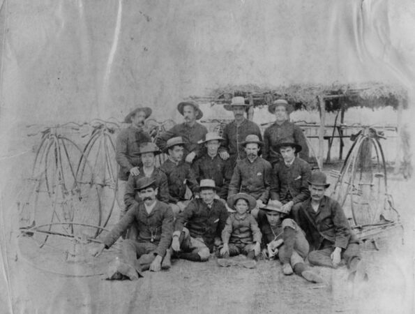 Penny-farthing riders posing with their bicycles surrounding them. Possibly a cycling club.  The name penny-farthing refers to British penny and farthing coins, one much larger than the other so that the side view resembles a penny leading a farthing. (Information taken from Herlihy, David V., 2004, Bicycle, The History. Yale University Press.)