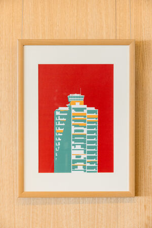 "Hot Modernism" poster in a frame on a wall, showing a modernist apartment building in red, yellow and teal