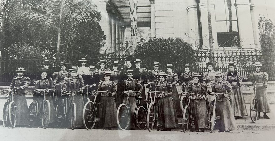 Black-and-white historical photograph of a large group of women posed with bicycles in front of a grand building with columns and a wrought-iron fence. The women wear long dresses and hats, standing in rows, suggesting a formal gathering or cycling club from the late 19th century.