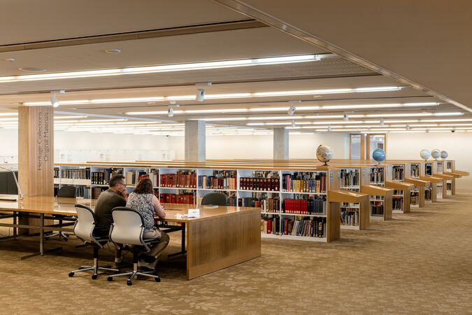 Interior of a library with rows of bookshelves. Two people are working together at a table.