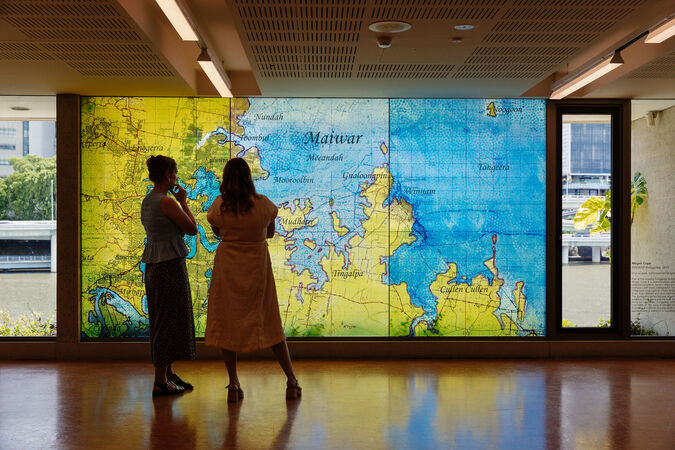 Two woman stand in front of the art installation "MAIWAR Yunggulba" by Megan Cope; it is a large topographical map on a glass window