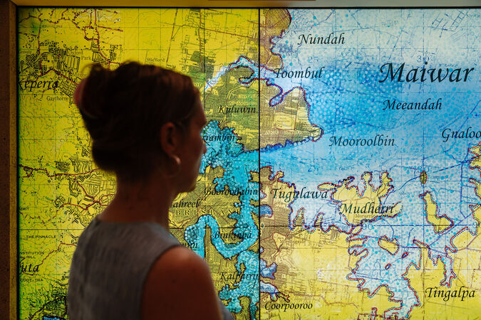 A woman stands in front of the art installation "MAIWAR Yunggulba" by Megan Cope; it is a large topographical map on a glass window