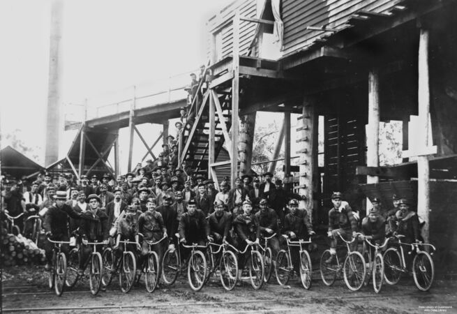 Black-and-white photograph of a large group of miners gathered outside an industrial timber structure, many seated on bicycles in the front row. The men wear work clothes and hats, some with lamps, suggesting a coal mining community. Additional workers stand on stairs and platforms behind them.