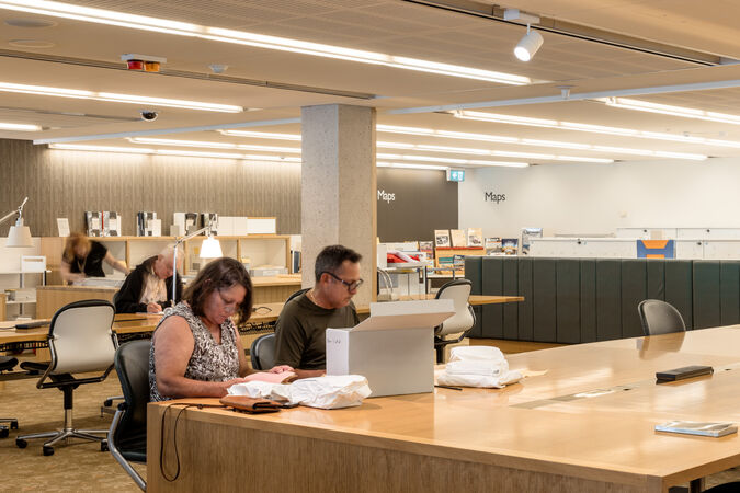 Patrons at a library sit at desks looking through research material. In the background is a section for maps.