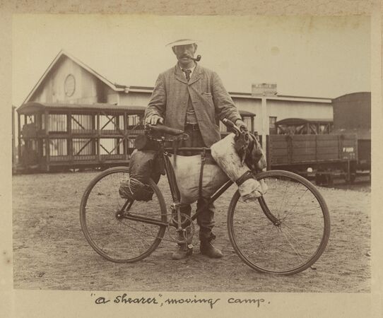 Sepia-toned photograph of a man standing with a heavily loaded bicycle, carrying bags and equipment. He wears a hat and jacket and smokes a pipe. Behind him are timber buildings and a rail wagon, suggesting a rural worksite. Caption reads, “a shearer, moving camp.”