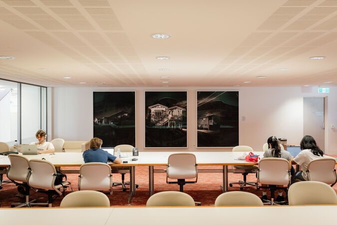 Four people sit at desks in the library; in the background is the artwork 'Sixteen – Triptych' by Ian Strange