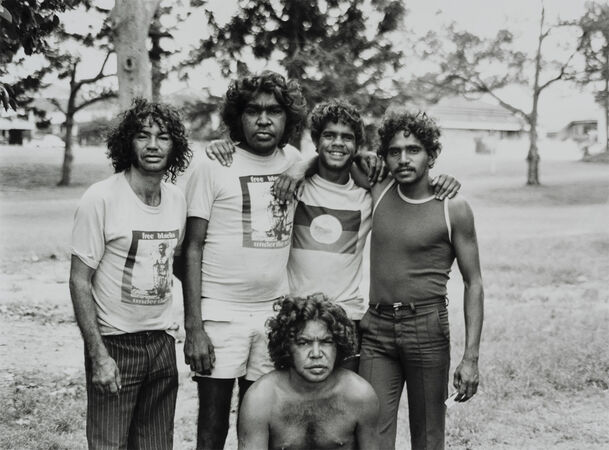 Group portrait of five young Aboriginal men outdoors, standing and crouching together with arms around each other.