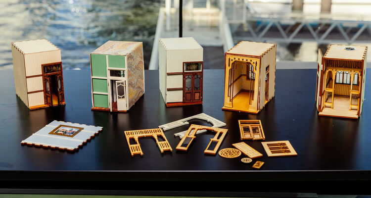 A row of detailed miniature house models made from wood and paper sits on a black table with various decorative parts laid out in front of them. The background shows a waterfront scene through a large window.