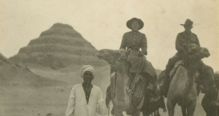 Two people on camels being led by two men with step pyramid in background