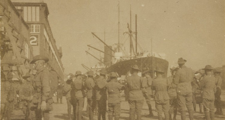 Faded picture of a group of men in uniform facing away from the camera toward a large ship, with buildings to the left.