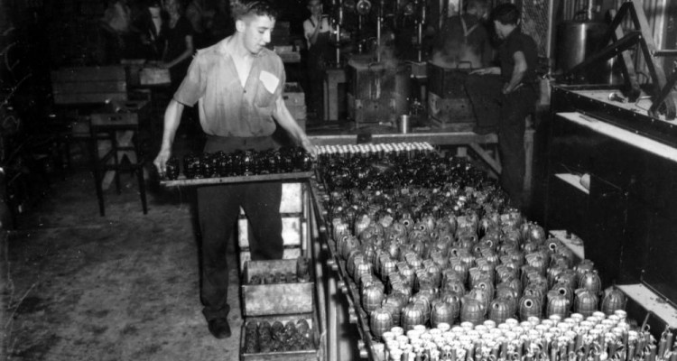Black and white phorograph of a man in a factory, stacking crates of grenages.