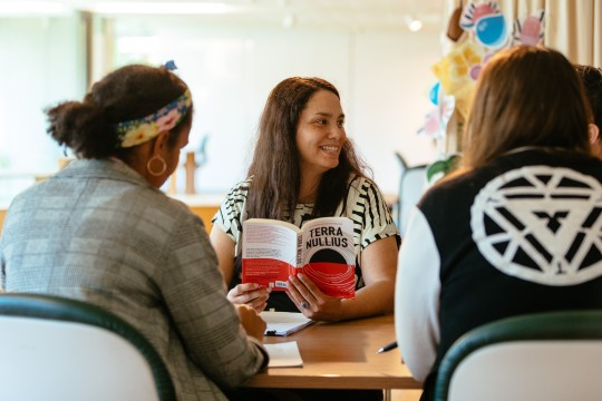 Three people are sitting at a table. The one person facing the camera is holding the book 'Terra Nullius' and is smiling to the side. 