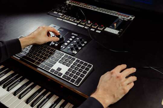 a close up shot of a man's hands that are using the recording studio at the edge. One hand is on a computer mouse the other is adjusting an audio dial. A keyboard is visible in the bottom corner. 