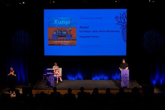 one person stands behind a lectern on stage speaking and another sits on-stage listening. There is an Auslan interpreter standing to the side of the stage.