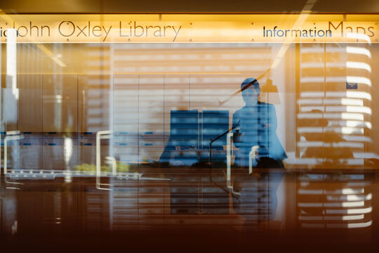 State Library staff member behind information desk. Reflective glass is in front of them.