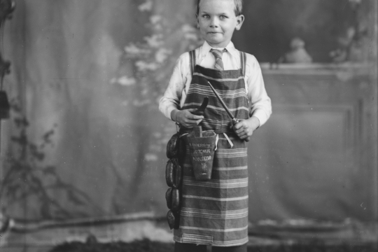 A black and white photograph of a young boy wearing a butchers apron and holding a string of sausages 
