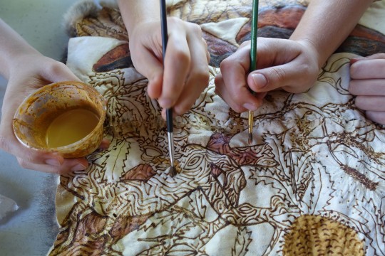 Two artists painting a possum skin cloak with ochre.