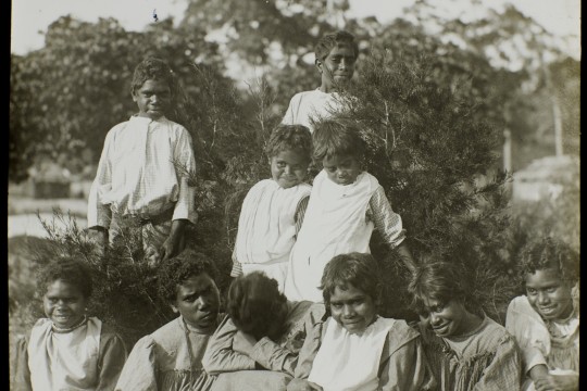 Black and white image of Aboriginal children, six sitting, three standing behind the others, in front of some bushes