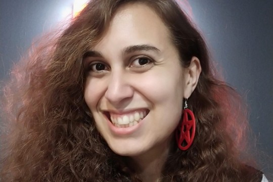 A photograph of writer Allanah Hunt, who has wavy brown hair haloed by soft lighting and wears red pentagram earrings