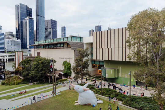 A wide view of State Library of Queensland from GOMA, showing an inflated silk bilby on Maiwar Green with children playing near it.