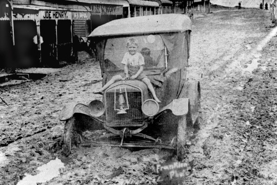 Young boy sitting on the bonnet of an early motor vehicle submerged in mud in Rankin Street, Innisfail in 1925. The street is covered in mud and water from the flood which hit the town in March 1925. The car is a Ford Model T between 1917-1927.