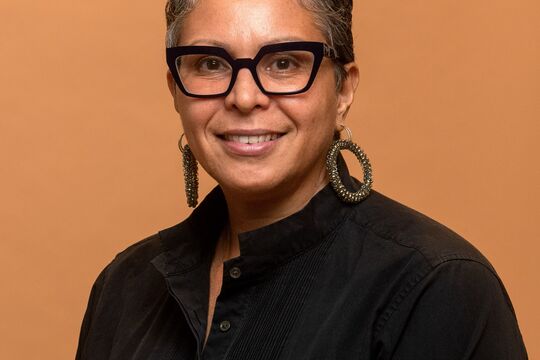 Photo of Cheryl Leavy against an orange background. Cheryl wears glasses and a black shirt.