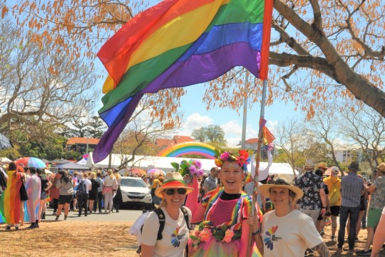 Three women standing together in a park holding a rainbow flag with crowd in background