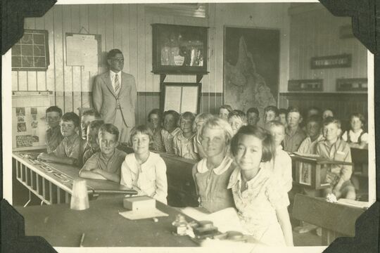 4 rows of children sitting in front of desks in a classroom with adult male standing at back of classroom