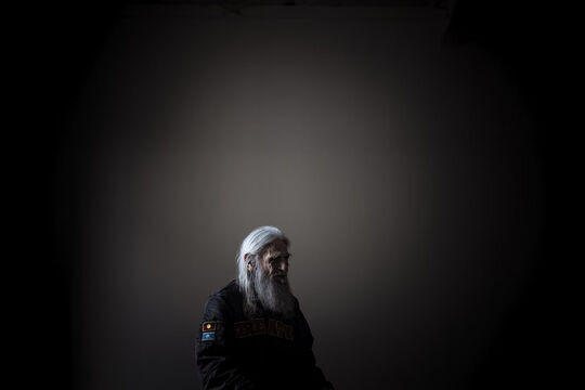 An indigenous elder sits in a dark room with a vignetted light on him. 