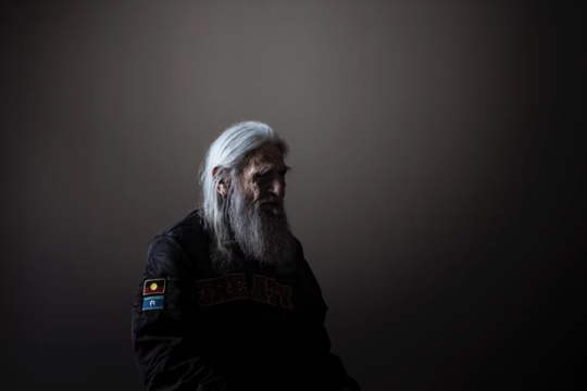 An older man with long white hair and beard sits in an empty room with a vignette of light on him. 