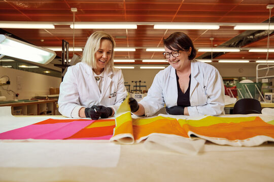 Two women wearing white lab coats and gloves carefully handling and preparing a fabric roll for preservation storage.