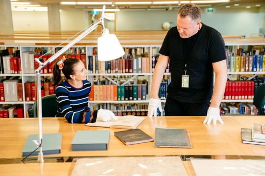 Visitor talking to State Library staff member. The visitor is wearing white gloves while pointing to old books.