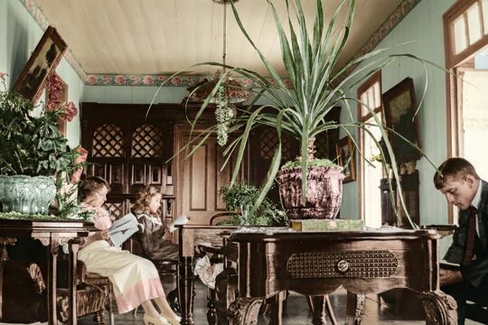 A colourised early 20th-century interior showing three children and a man reading in a well-furnished room, with plants, wooden furniture, and soft natural light creating a calm atmosphere.