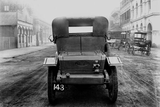 Black-and-white photograph showing the rear of an early motor car parked in the middle of a wide, unsealed street, with horse-drawn carts and colonial-era buildings lining both sides of the road