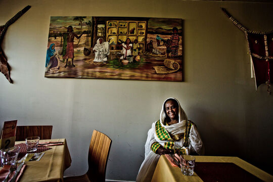 Woman seated in a restaurant, with a table beside her and a painting on the wall behind.