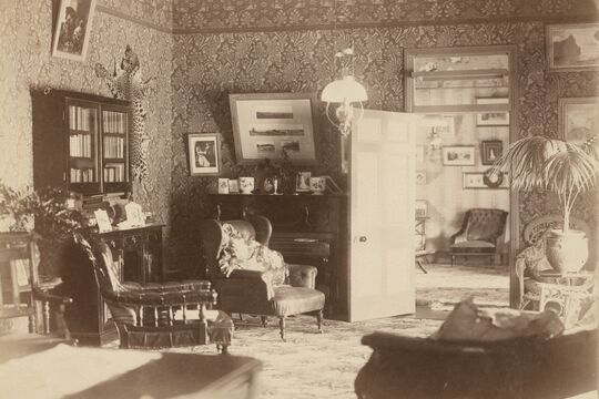 A sepia-toned photograph of a well-furnished early 20th-century living room featuring patterned wallpaper, upholstered armchairs, a piano with framed photographs on top, a glass-front bookcase, and decorative items including a mounted animal skin.
