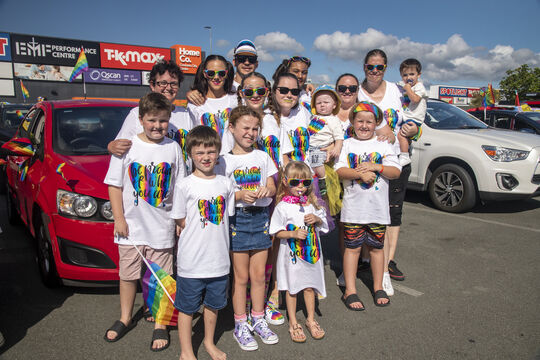 A group of adults and children wearing rainbow t-shirts standing together in front of a red car.