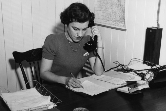 Black and white image of woman sitting at a desk writing while holding a telephone to her ear, ca 1952