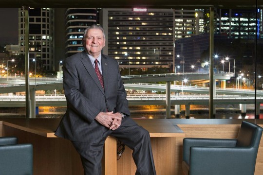 James C Sourris, a smiling man in a suit sits in the library with his back to the Brisbane cityscape at night.