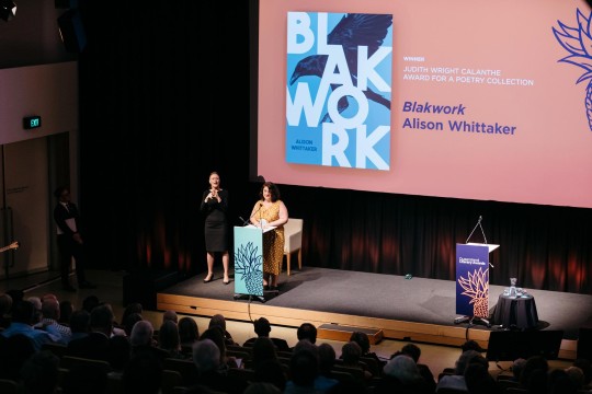 Person speaking at a lectern with a large projection behind her. An Auslan interpreter is beside her.