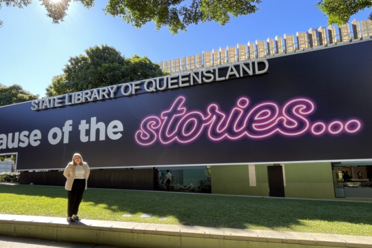 Photo of Kelly Whiteway standing out the front of the State Library of Queensland in front of the main signage board, which reads State Library of Queensland, Because of the stories